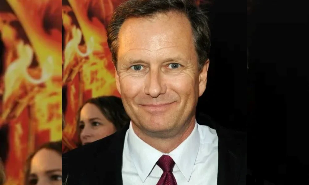 Jennifer Landon Husband at a public event, wearing a dark suit and maroon tie, standing in front of a fiery background poster.