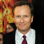 Jennifer Landon Husband at a public event, wearing a dark suit and maroon tie, standing in front of a fiery background poster.