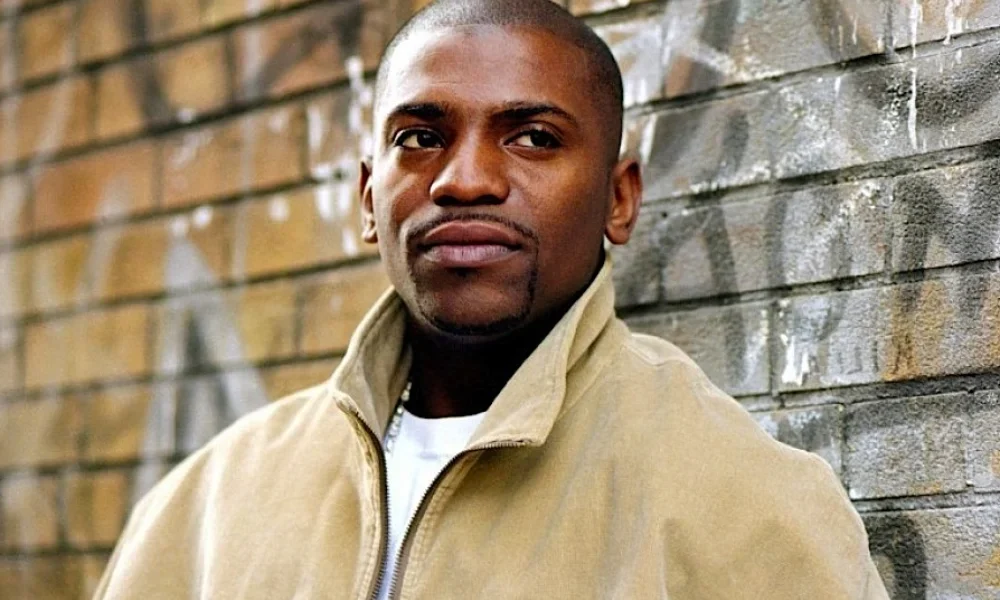 Omikaye Phifer standing in front of a graffiti-marked brick wall, wearing a beige jacket and displaying a confident, thoughtful expression