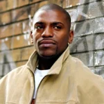 Omikaye Phifer standing in front of a graffiti-marked brick wall, wearing a beige jacket and displaying a confident, thoughtful expression